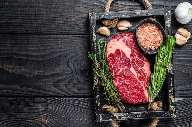 Bistecca di carne di manzo prime costolette crude in un vassoio di legno del macellaio con erbe sulla tavola di legno. Vista dall'alto.