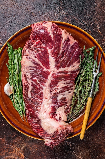 Bistecca di carne di manzo tenera o singola d'attaccatura cruda sul piatto rustico con le erbe. Sfondo scuro. Vista dall'alto.