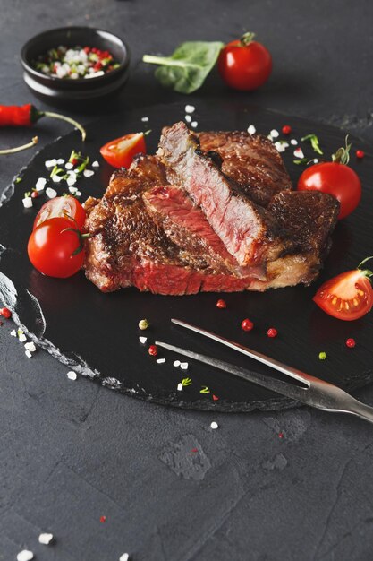 Bistecca di costata di manzo rara su piatto scuro decorato con pomodori, peperoncino, spezie e forchetta per carne, spazio copia