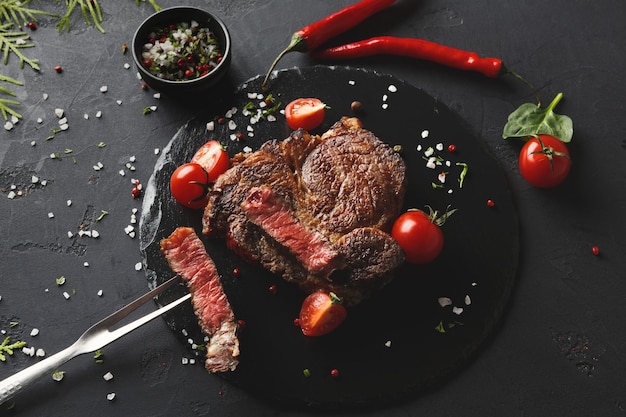 Bistecca di costata rara su piatto scuro decorato con pomodori, peperoncino, spezie e forchetta per carne, vista dall'alto, spazio copia