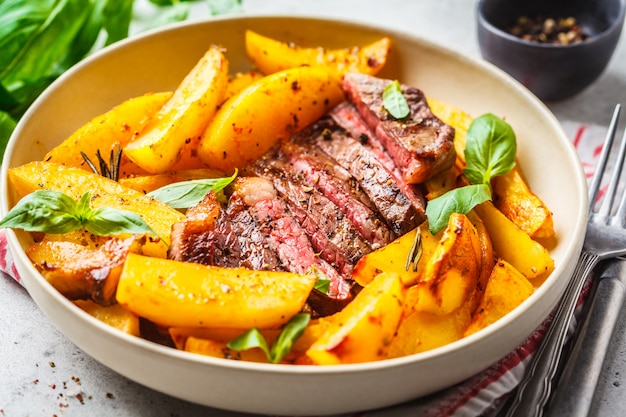 Bistecca di manzo alla griglia con patate e basilico in un piatto bianco su sfondo bianco.