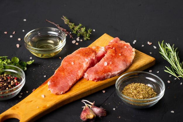 Bistecche di carne cruda su un tagliere Sfondo scuro Preparazione per la cottura della carne di maiale Vari condimenti di spezie si trovano nelle vicinanze