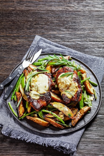 Bistecche di maiale condite con formaggio blu fuso e rosmarino su un pad di zucchero arrosto piselli spicchi di patate fagiolini e aglio su un piatto nero vista verticale