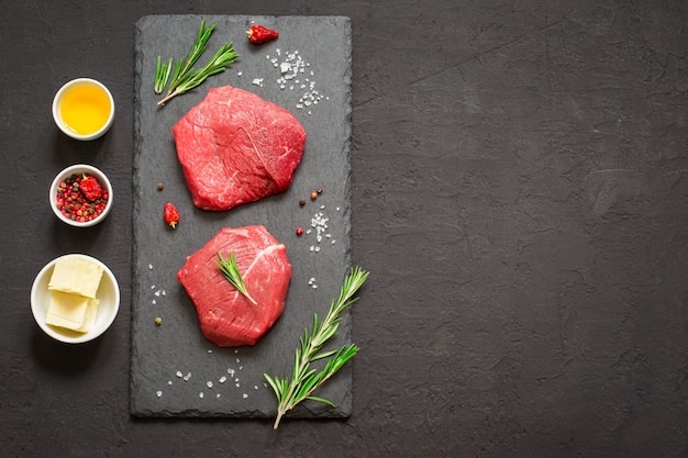 Bistecche di manzo di carne cruda su tavola di ardesia nera, spezie e ingredienti per cucinare. Vista dall'alto con copia spazio