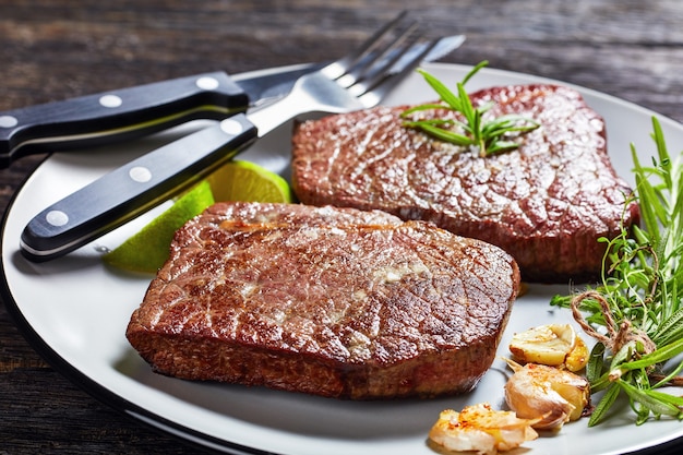 Bistecche di manzo fritte con rosmarino, aglio e lime su una piastra con forchetta e coltello su un tavolo di legno scuro, vista orizzontale dall'alto, primo piano