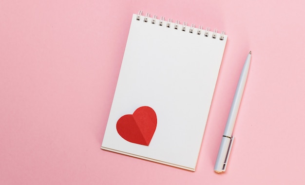 Blocco note in bianco, penna e cuori di carta rossa su sfondo rosa. Messaggio d'amore. San Valentino e concetto di vacanza romantica. Vista dall'alto, piatto con spazio di copia.