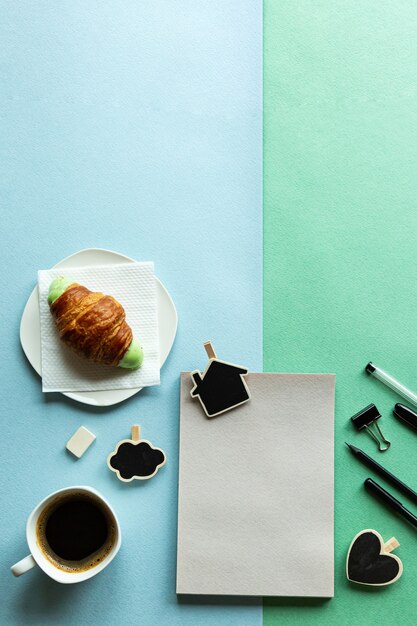 Blocco note, matita, croissant e caffè su una scrivania verde-blu.