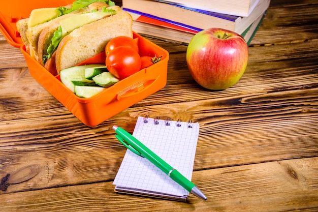 Blocco note penna mela matura pila di libri e pranzo al sacco con cetrioli panini e pomodori sul tavolo di legno