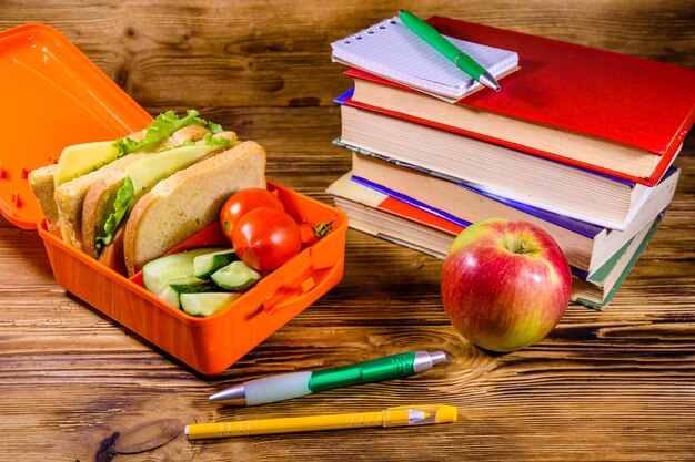 Blocco note penne mela matura pila di libri e pranzo al sacco con panini cetrioli e pomodori su tavola di legno rustico