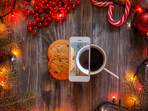 Blog e concetto di fotografia mobile. decorazioni festive di natale e capodanno su sfondo di legno, lucine e stringhe di perline che circondano una tazza di caffè e biscotti con gocce di cioccolato.