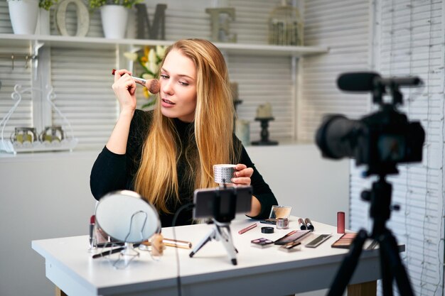 Blogger di bellezza donna che filma la routine quotidiana di trucco tutorial alla telecamera su treppiede Influencer girl live streaming cosmetica recensione del prodotto in home studio Vlogger donna applica la polvere per la pelle con pennello