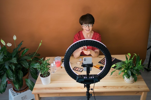 Blogger di bellezza femminile di mezza età o influencer con prodotti per il trucco che registrano video nella stanza di casa Utilizzando lampada ad anello e smartphone