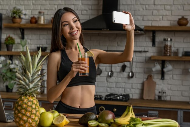 Blogger femminile sportivo che fa selfie con un frullato utilizzando lo smartphone nella cucina di casa