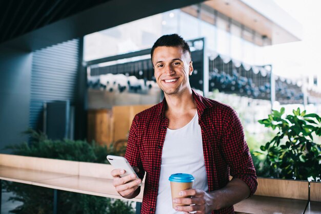 Blogger maschio con tazza da asporto e moderno dispositivo cellulare che sorride alla telecamera