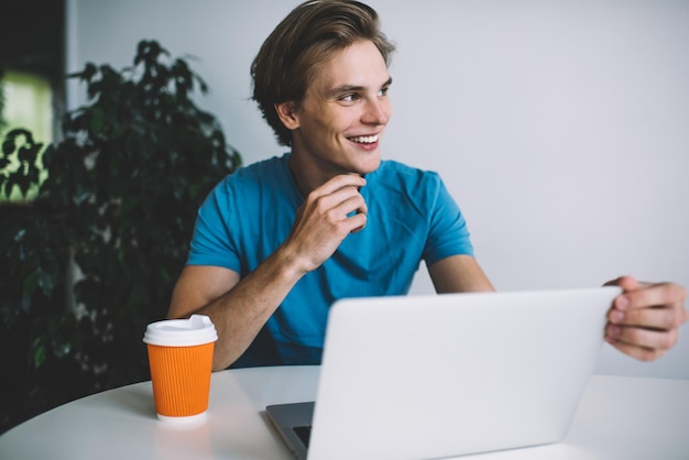 blogger maschio con un sorriso carino sul viso che distoglie lo sguardo e si rallegra del tempo libero per il networking tramite laptop