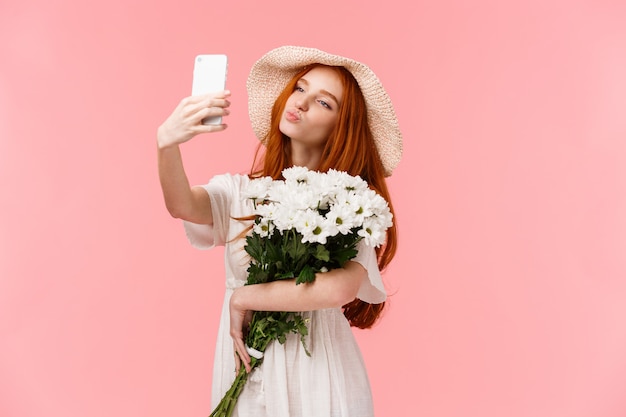 Blogger sciocca e carina, femminile che prende selfie con il bouquet di fiori portato dal ragazzo, che tiene smartphone che piega le labbra nel bacio dell'aria, posa per uno scatto sensuale perfetto, stand rosa