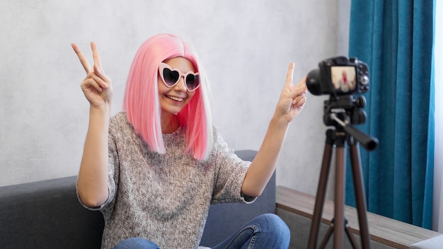 Blogger teenager felice della ragazza con il fronte sorridente in parrucche e occhiali rosa. Mostra il segno della vittoria, guardando la telecamera che registra un vlog in diretta, facendo una videochiamata