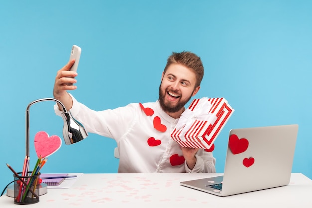 Blogger uomo allegro positivo con barba in camicia bianca ricoperta di adesivi a forma di cuore che tengono e mostrano sulla fotocamera dello smartphone confezione regalo a strisce Colpo in studio al coperto isolato su sfondo blu