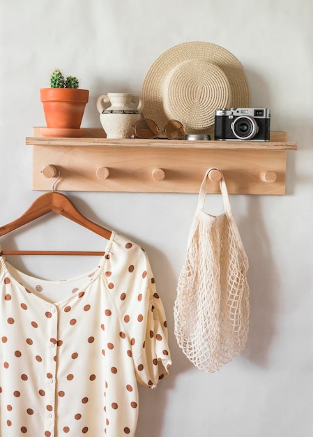 Blusa estiva da donna con pois su un appendiabiti su una mensola di legno con un cappello di paglia una borsa ecologica una macchina fotografica in una stanza luminosa Atmosfera di vacanza