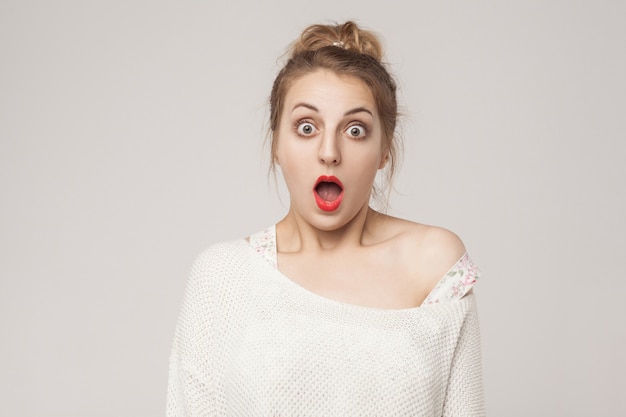 Bocca aperta bionda di stupore e urla con faccia sorpresa. Foto in studio, sfondo grigio