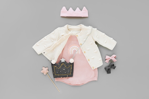 Body rosa con maglione lavorato a maglia, borsa per bambini, corona in cotone e bacchetta magica. Set di vestiti e accessori per bambini su sfondo grigio. Vestito alla moda per bambini. Disposizione piatta, vista dall'alto