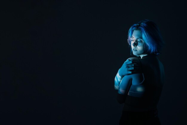 Body shaming Ritratto d'arte Autoaccettazione Imbarazzata timida donna imperfetta con gli occhiali che si abbraccia alla luce al neon blu isolata su uno sfondo di spazio vuoto notte oscura