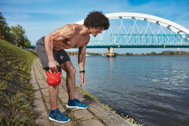 Bodybuilder con six pack, ABS perfetto, spalle, bicipiti, tricipiti e petto sta facendo un allenamento del corpo in forma forte con kettlebell, vicino al fiume.