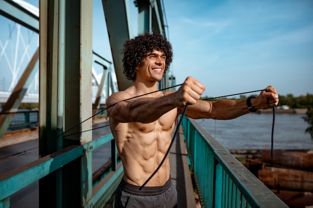 Bodybuilder con torso nudo, sta facendo un allenamento del corpo in forma forte con elastico, sul ponte del fiume.