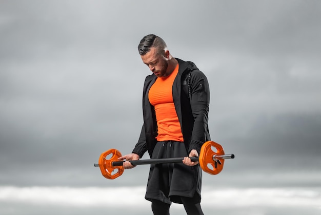 Bodybuilder in abbigliamento sportivo arancione che fa esercizi con il barbell per i bicipi in una palestra all'aperto potente