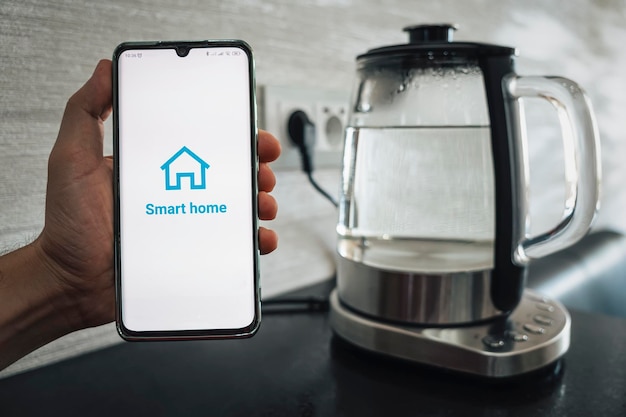 Bollitore elettrico trasparente con acqua bollente in cucina uno smartphone in mano a un uomo per il controllo remoto di un bollitore intelligente