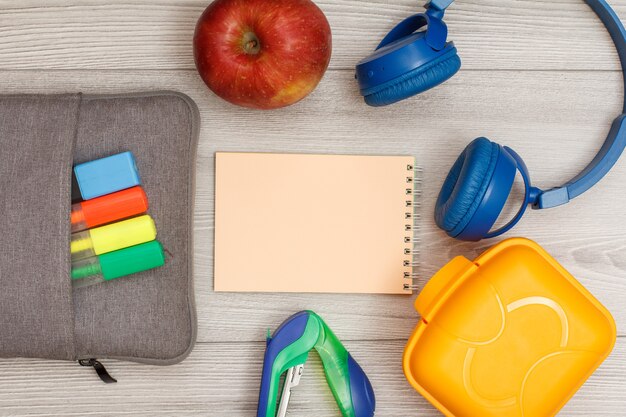 Borsa-astuccio con pennarelli colorati e pennarello, mela, quaderno rosa, cucitrice, cuffie e scatola per il pranzo su fondo di legno grigio. Vista dall'alto con copia spazio. Torna al concetto di scuola. Materiale scolastico.