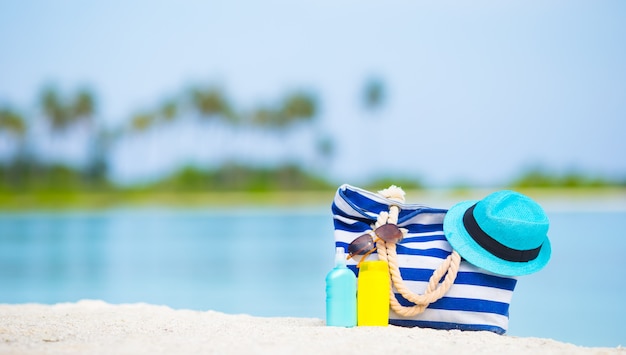 Borsa blu, cappello di paglia, occhiali da sole e bottiglie di crema solare sulla spiaggia bianca