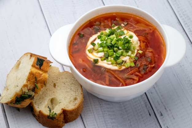 Borscht rosso ucraino con panna acida in ciotola bianca e pezzi di pane con aglio. Concetto di cucina russa. Tavole di legno chiaro sfondo, tavolo.