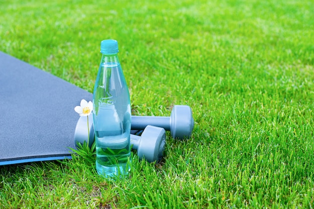 Bottiglia con acqua, manubri e una stuoia per lo sport e lo yoga sull'erba verde. Copia spazio