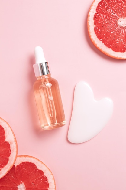 Bottiglia con contagocce di olio essenziale aromatico con fetta fresca di pompelmo Rullo facciale e guasha su sfondo rosa Cosmetici creativi alla moda piatto con spazio per la copia