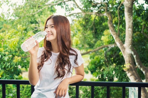 Bottiglia di acqua asiatica sorridente felice della tenuta della donna da bere, stante sul suo balcone