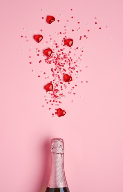 Bottiglia di champagne con spruzzata di coriandoli a forma di cuore rosso e cuori decorativi su sfondo rosa. Concetto di San Valentino. Vista dall'alto, copia dello spazio.