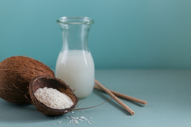 Bottiglia di latte vegano al cocco con cannucce, cocco intero e fiocchi su fondo azzurro