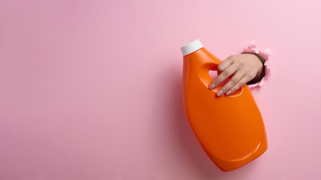 Bottiglia di plastica arancione con detersivo liquido in una mano femminile su sfondo rosa. Una parte del corpo sporge da un buco strappato sullo sfondo, un posto per un'iscrizione