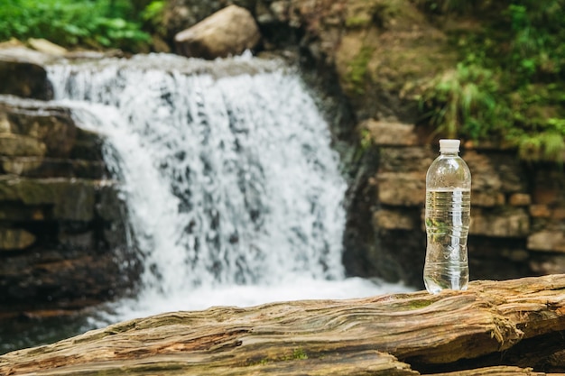 Bottiglia di plastica trasparente con acqua su un tronco di legno sullo sfondo di una cascata