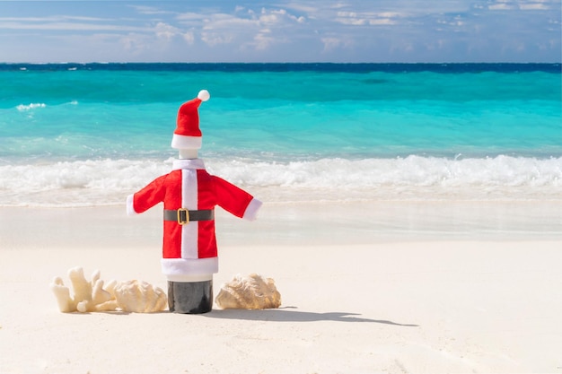 Bottiglia di vino decarata con cappello e cappotto di Babbo Natale su spiaggia esotica e oceano blu nessuno