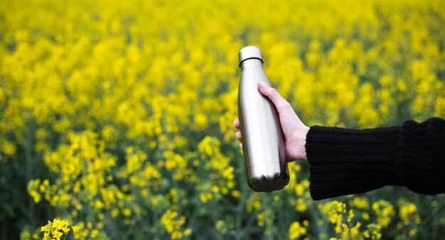 Bottiglia thermos inossidabile in mano, sulla superficie del campo di colza.