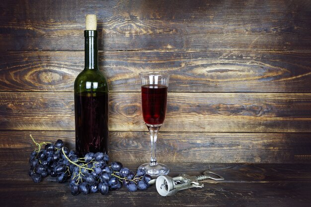 Bottiglia verde scuro e bicchiere da vino trasparente con vino rosso e grappolo d'uva sulla superficie della plancia di legno marrone, cavatappi nelle vicinanze