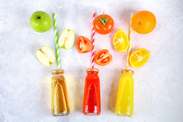 Bottiglie con arancia fresca, mela, succo di pomodoro e tubuli colorati su un tavolo di cemento grigio.