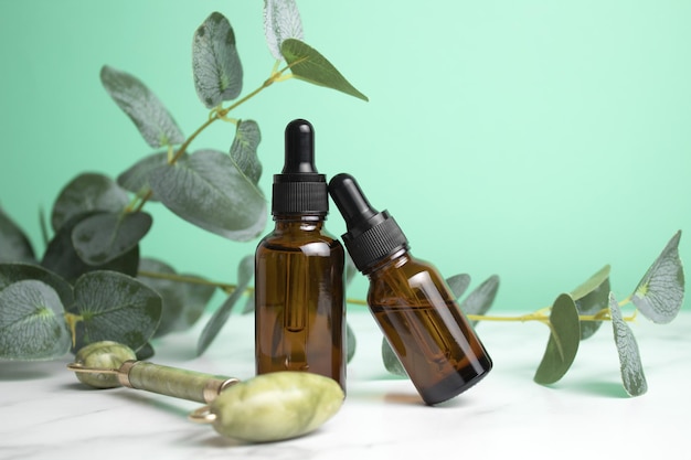 Bottiglie di vetro con rullo facciale in giada d'olio per massaggio facciale su sfondo verde con foglie di eucalipto