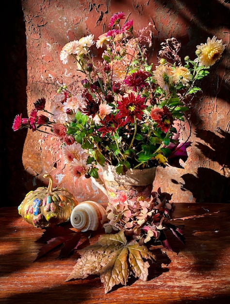 Bouquet autunnale e natura morta romantica. Un bouquet con fiori autunnali in un vaso di ceramica, zucca dipinta e conchiglia sotto i raggi del sole autunnale.