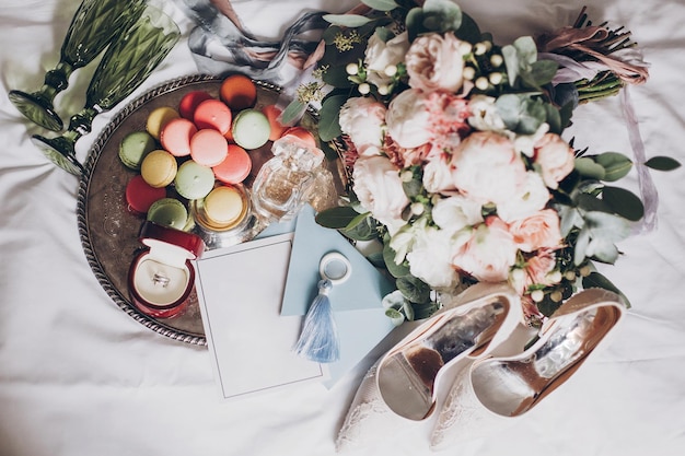 Bouquet da sposa moderno fedi nuziali elegante invito bottiglia di profumo scarpe deliziosi macarons e bicchieri verdi per champagne sul letto bianco Preparazioni nuziali o addio al nubilato