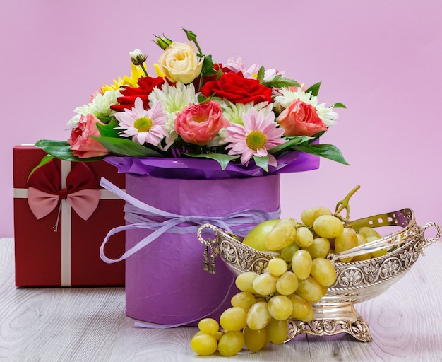 Bouquet di fiori di campo, confezione regalo e uva in un vaso di metallo su assi di legno.