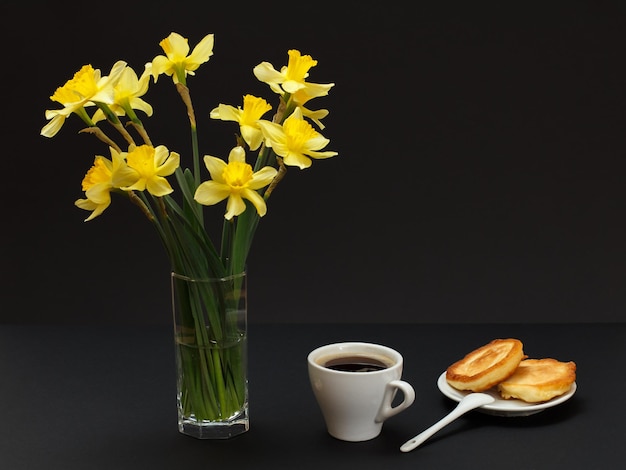 Bouquet di narcisi gialli in un vaso di vetro una tazza di caffè e un piatto con le frittelle