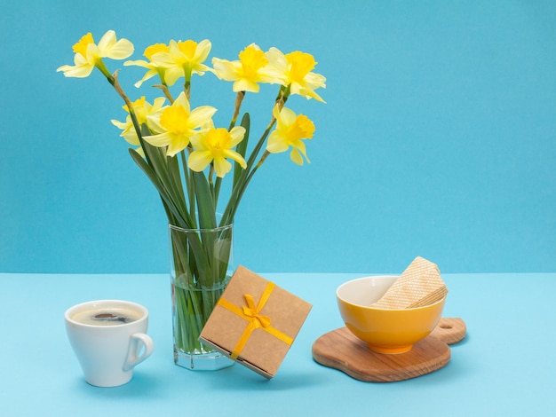 Bouquet di narcisi gialli in vaso di vetro una confezione regalo una tazza di caffè e un piatto con cialde